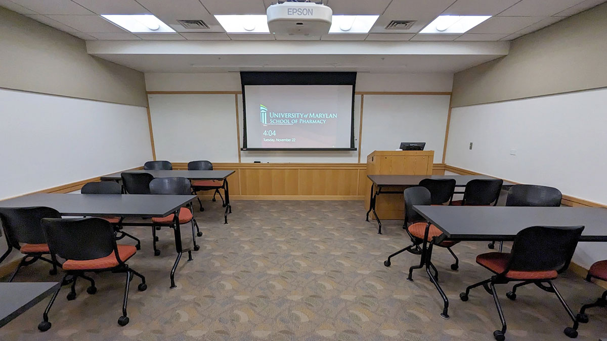 Small classroom with movable tables and chairs arranged in clusters, facing a projection screen and instructor podium.