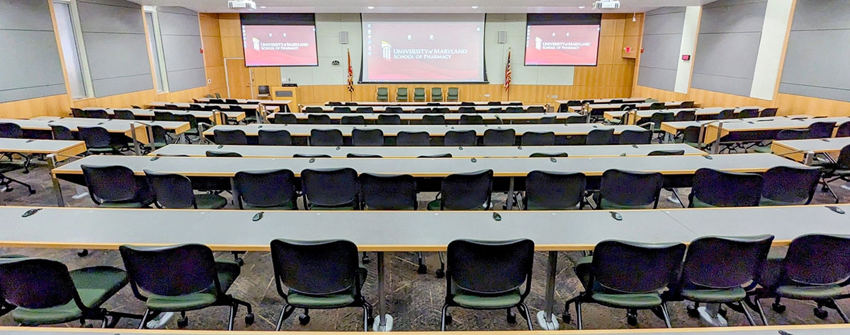 Wide view of an empty lecture hall in Pharmacy Hall North with tiered seating, long desks, and presentation screens at the front.