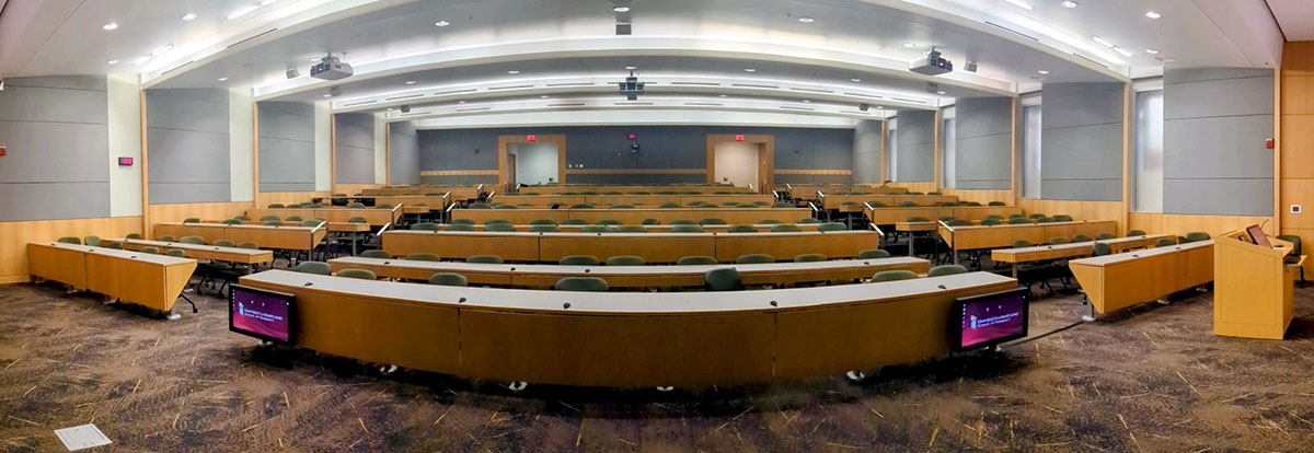 View from the front of an empty lecture hall in Pharmacy Hall North, showing curved, tiered rows of desks and chairs extending toward the back of the room.