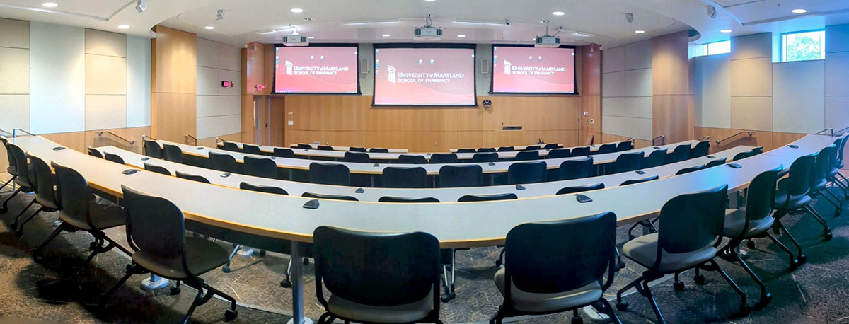 Medium‑sized lecture hall in Pharmacy Hall North with curved, tiered rows of desks and chairs facing multiple presentation screens.