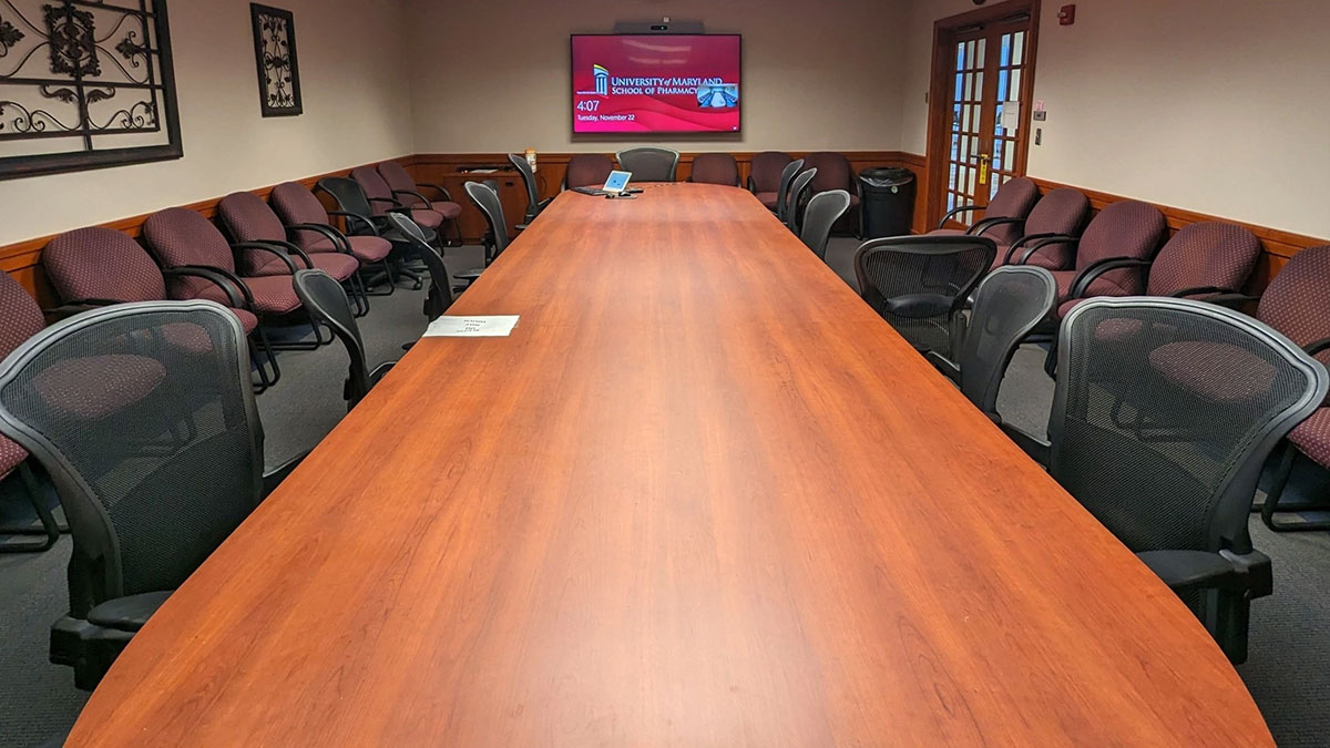 Conference room in Pharmacy Hall South with a long rectangular table, rolling chairs, additional seating along the walls, and a wall‑mounted display screen.