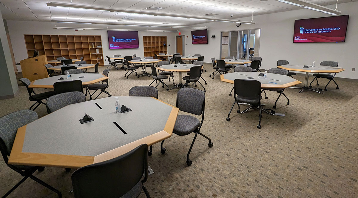 Patient interview area in Pharmacy Hall North Room N301A with multiple small tables and chairs arranged for interviews, wall‑mounted display screens, and open shelving along the back wall.