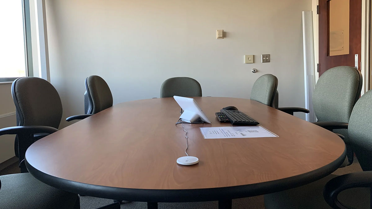 Alternate view of conference room Saratoga 01‑621 with an oval meeting table, rolling chairs, tabletop controls, and windows along one side.