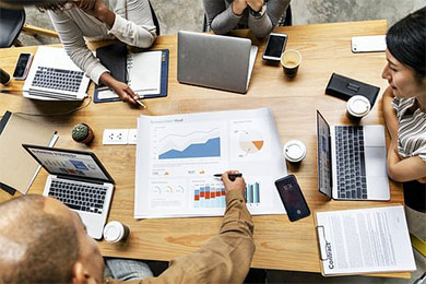 Professionals meet around a large table cluttered with laptops and spreadsheets.