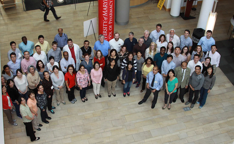 Large group of adults standing together in an indoor atrium, gathered in several rows and looking up toward the camera. A University of Maryland School of Pharmacy banner is visible behind them