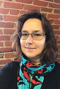 Kate Storm standing in front of a brick wall, wearing a colorful patterned scarf and dark shirt